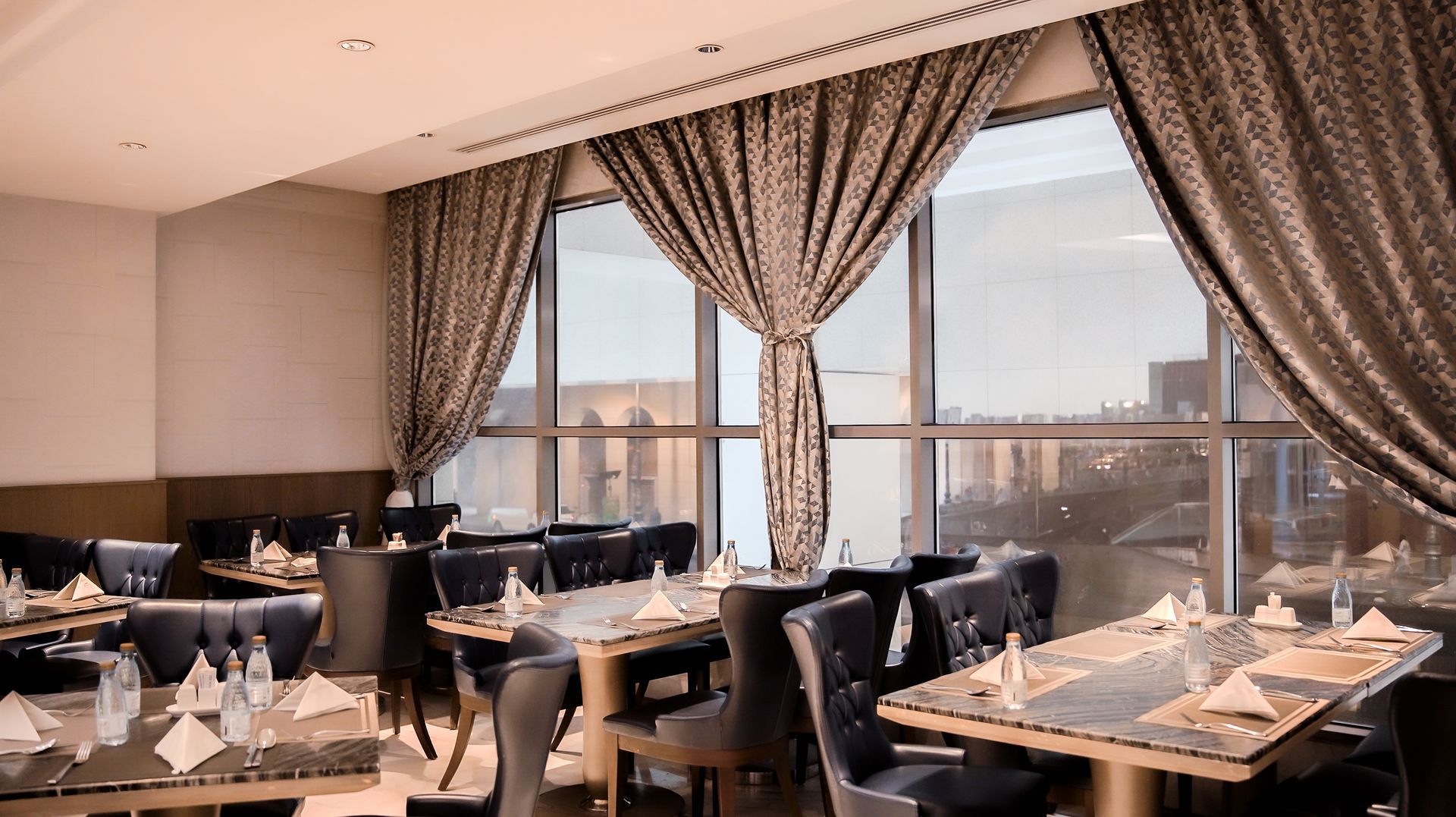 Elegant restaurant dining room with set tables, dark chairs, and large curtained windows overlooking the city