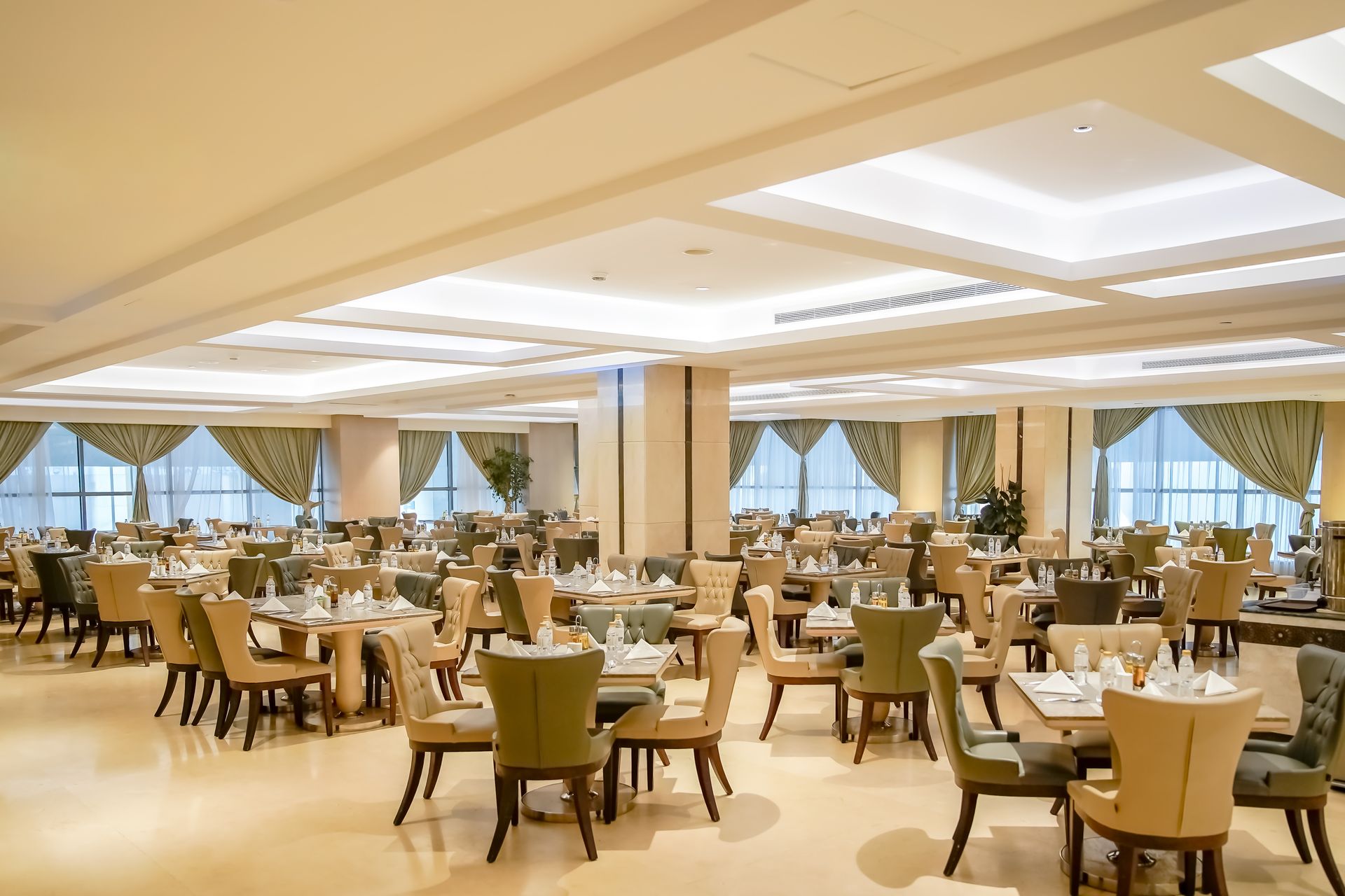 Elegant restaurant dining room with round tables, tan chairs, and large windows under soft ceiling lights