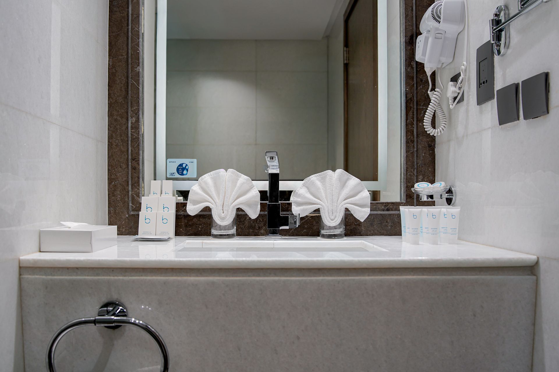 Hotel bathroom vanity with sink, mirror, folded towels, and toiletry items on the counter.