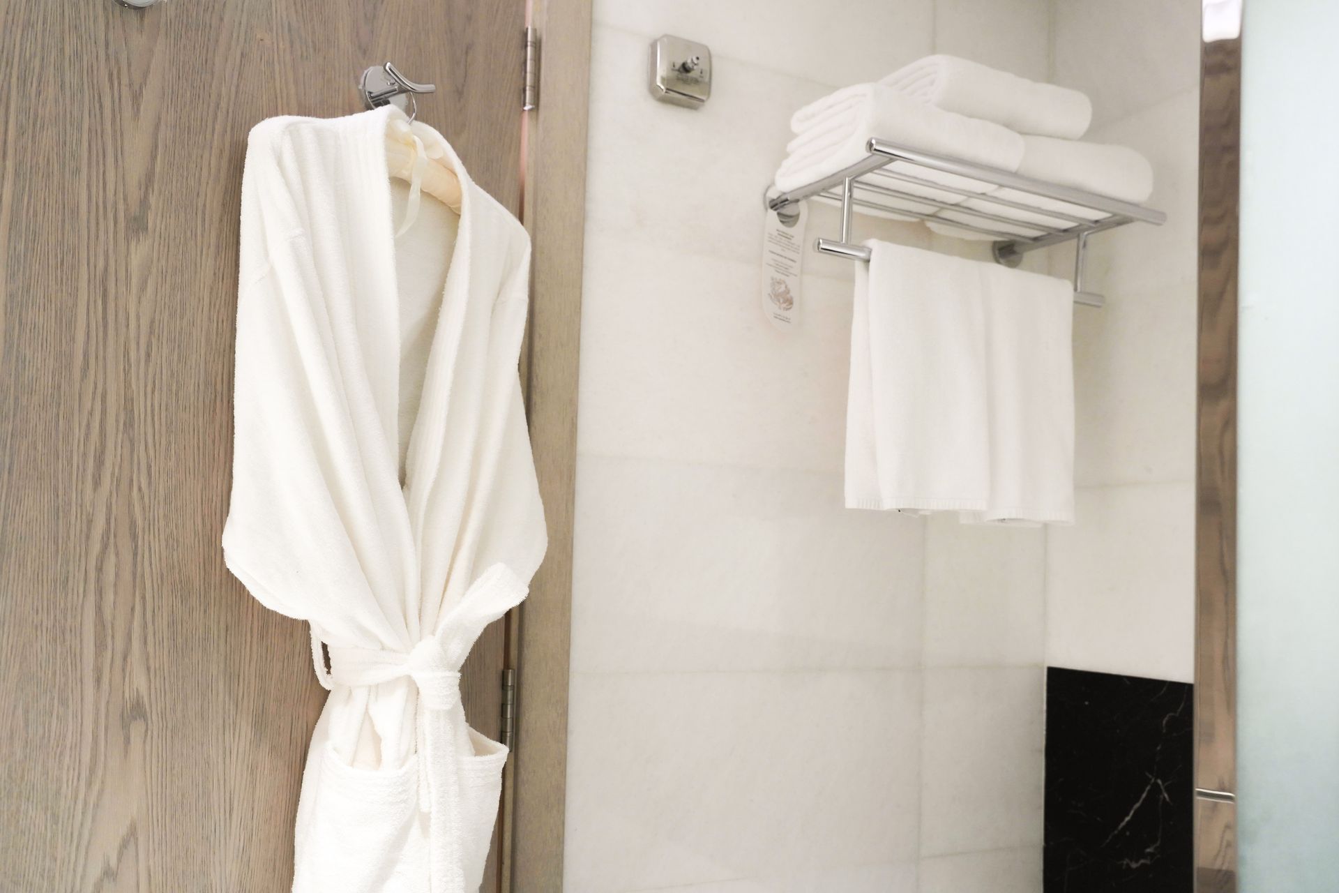 White bathrobes hang in a bathroom beside a towel rack with folded white towels.