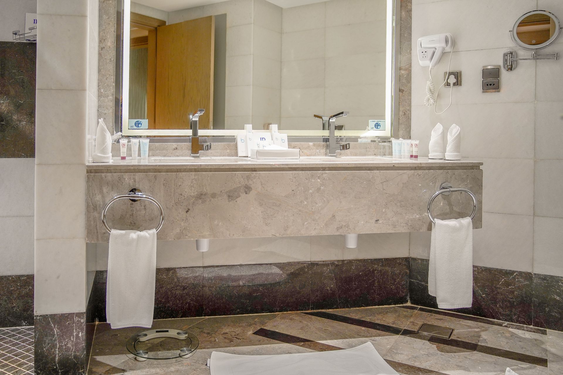 Modern bathroom sink with large mirror, wall sconces, and white towels on a marble vanity.
