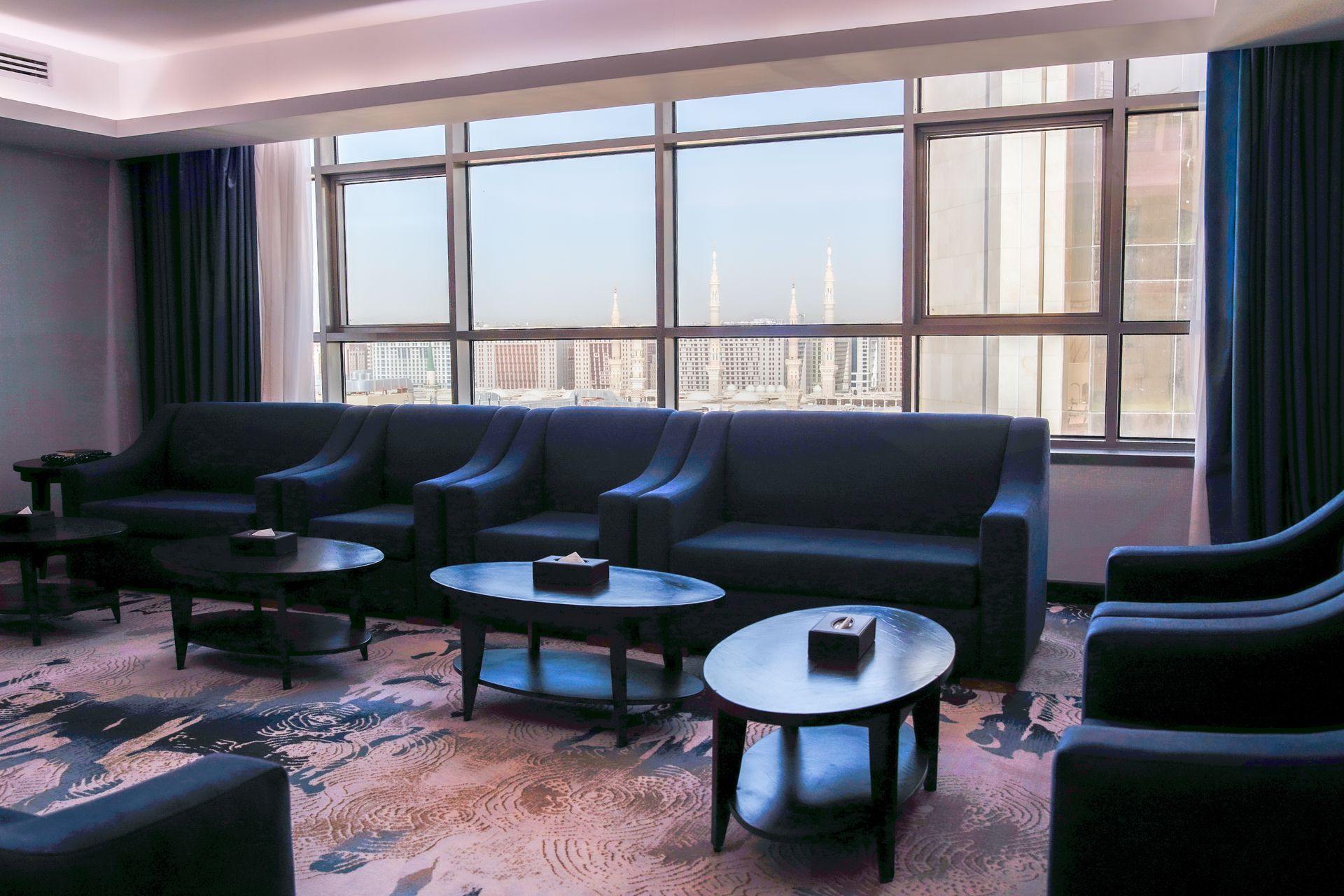 Modern living room with dark sofas, coffee tables, and large windows overlooking a city skyline