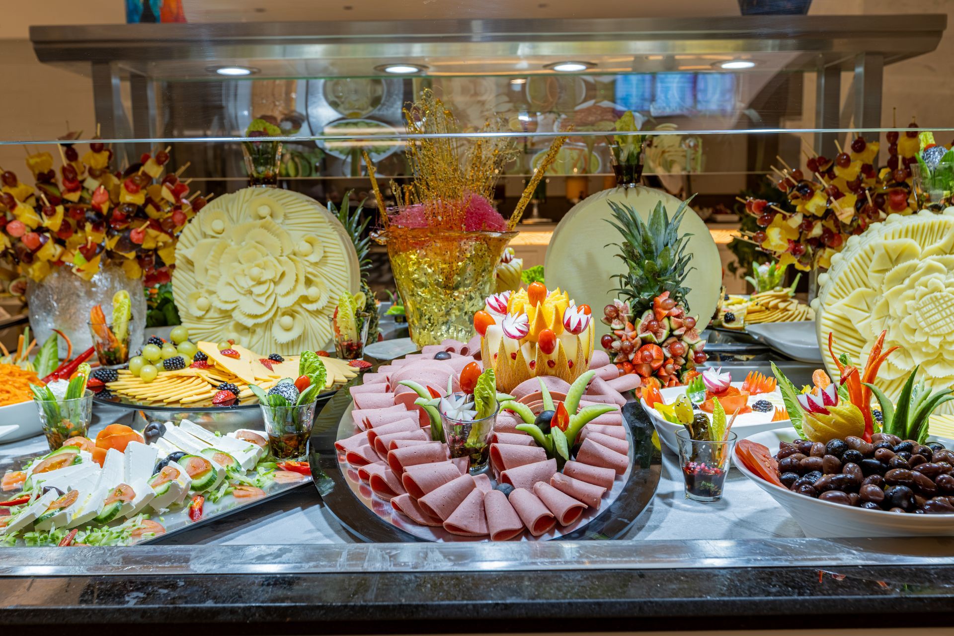 Colorful buffet display of sliced meats, salads, fruit, and garnishes under warm lights