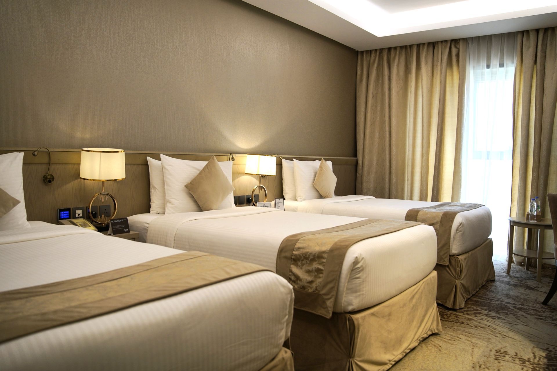 Hotel room with three neatly made beds, warm lighting, and a window with curtains