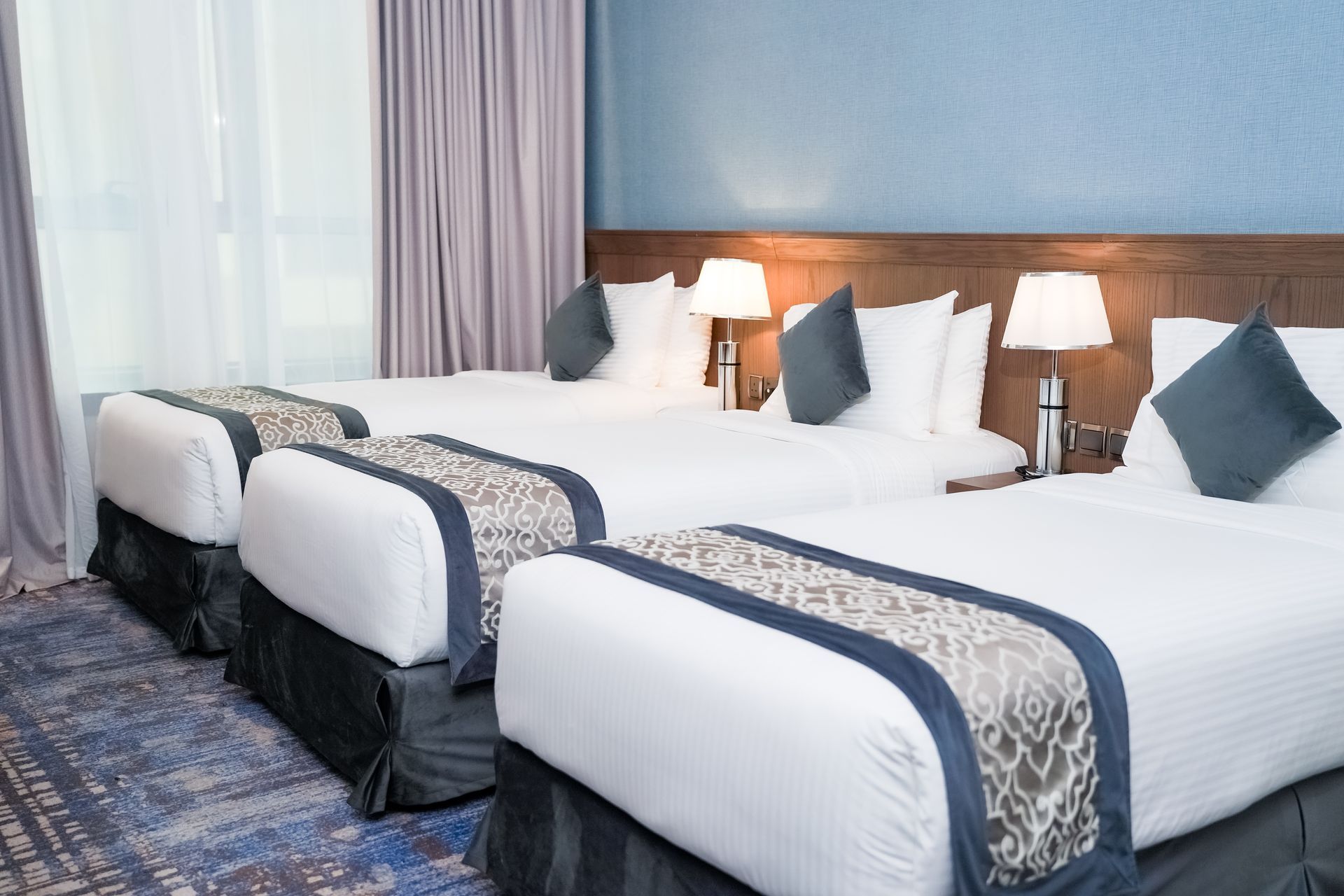 Hotel room with three neatly made twin beds, blue runners, bedside lamps, and a window with curtains