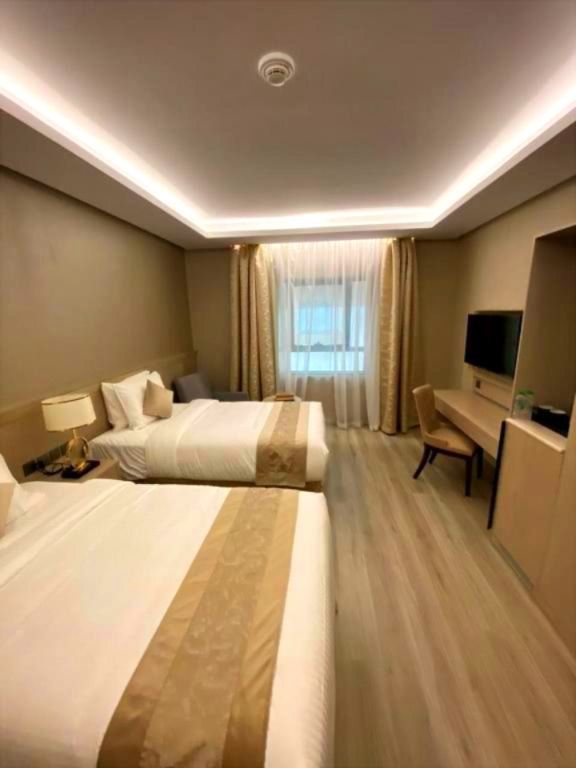 Modern hotel room with two beds, warm lighting, a desk, TV, and window with curtains.