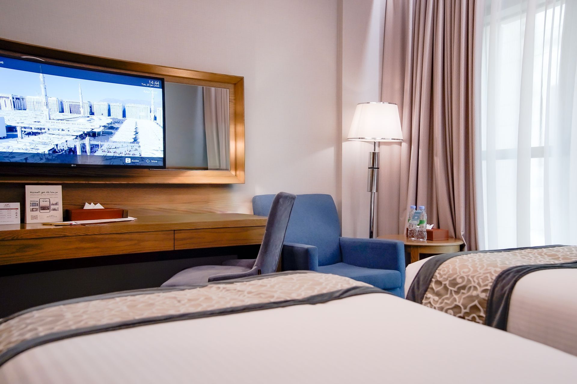 Modern hotel room with two beds, blue chair, wall-mounted TV, and lamp by a window with sheer curtains