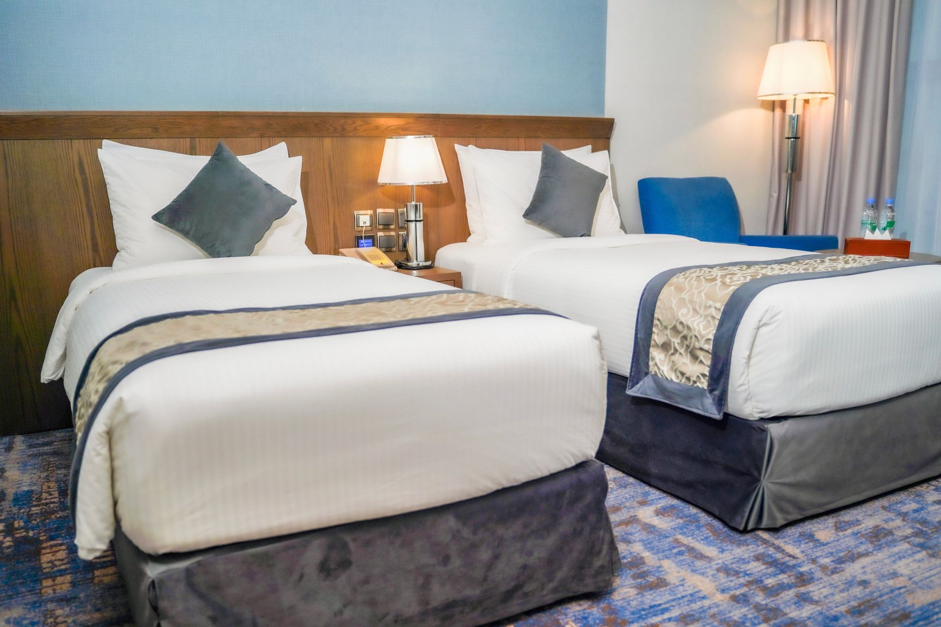 Hotel room with two neatly made beds, blue accents, a lamp, and a chair by the window.