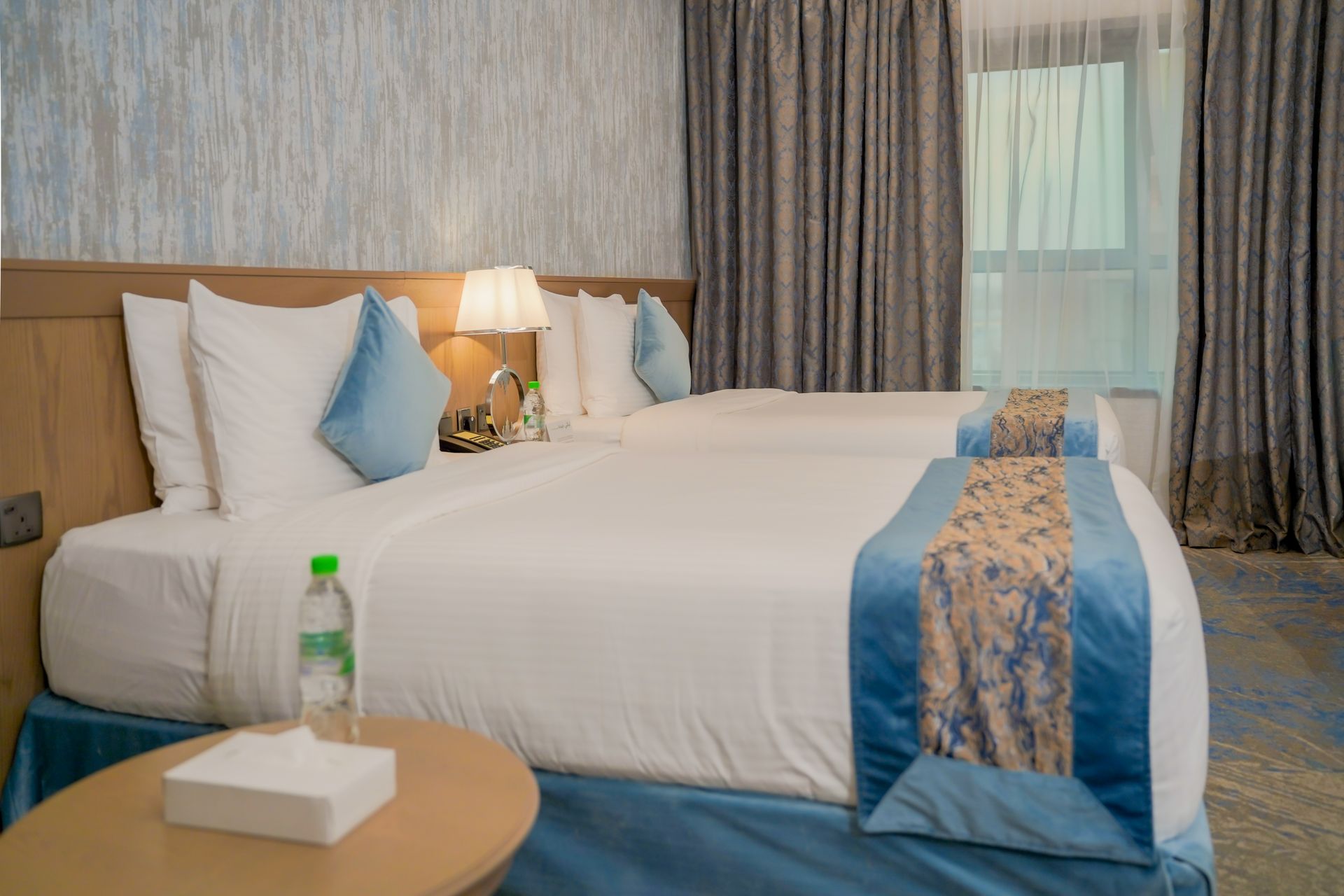 Modern hotel room with a neatly made bed, blue accents, bedside lamp, and curtains by the window