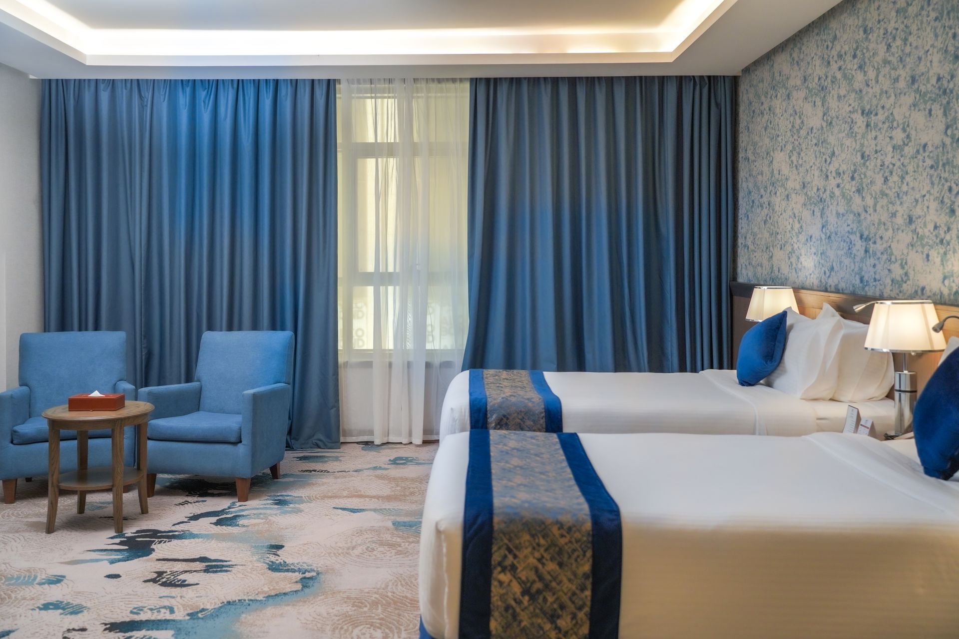 Blue hotel room with two beds, seating area, patterned carpet, and large curtains