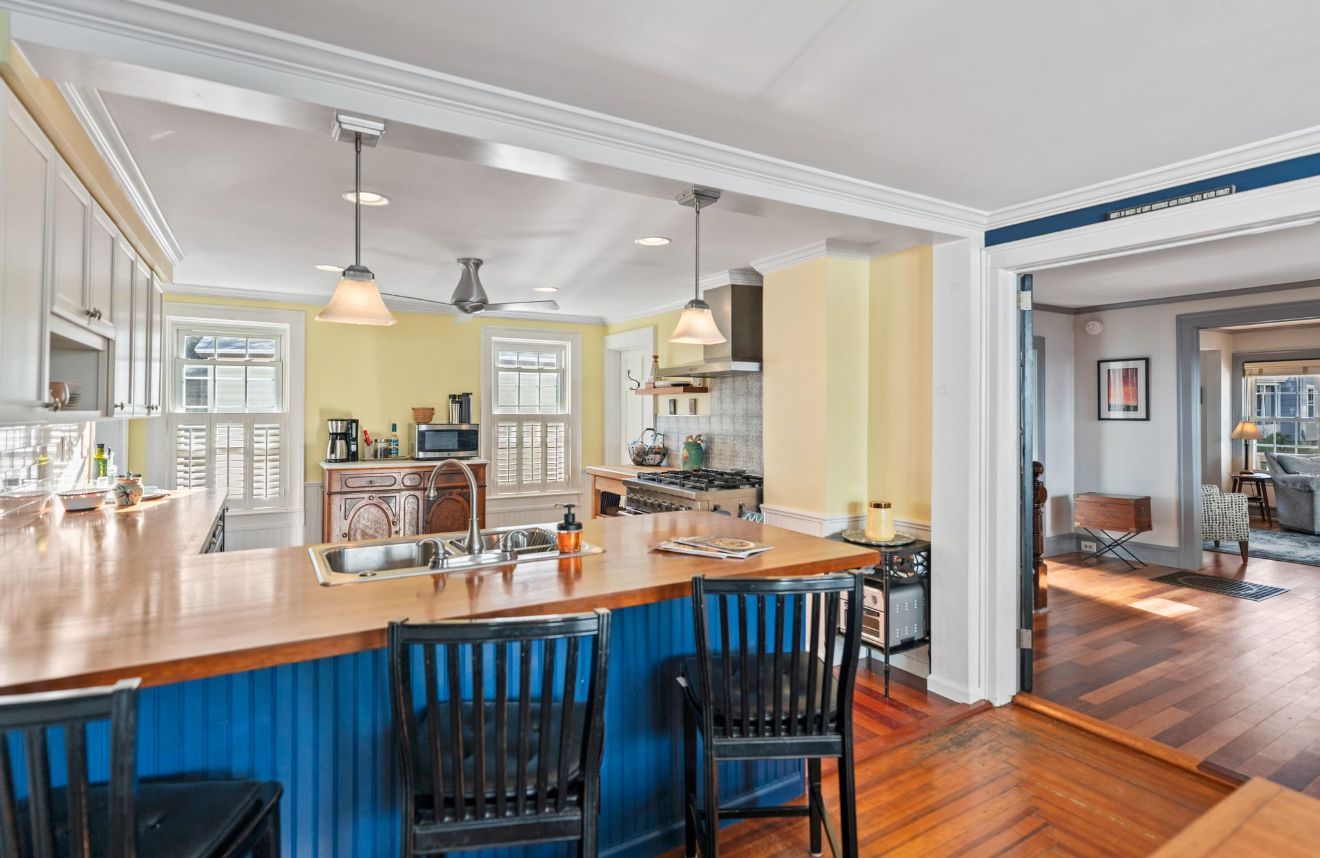 White porch with wicker chairs, overlooking a street with houses and trees under a blue sky.