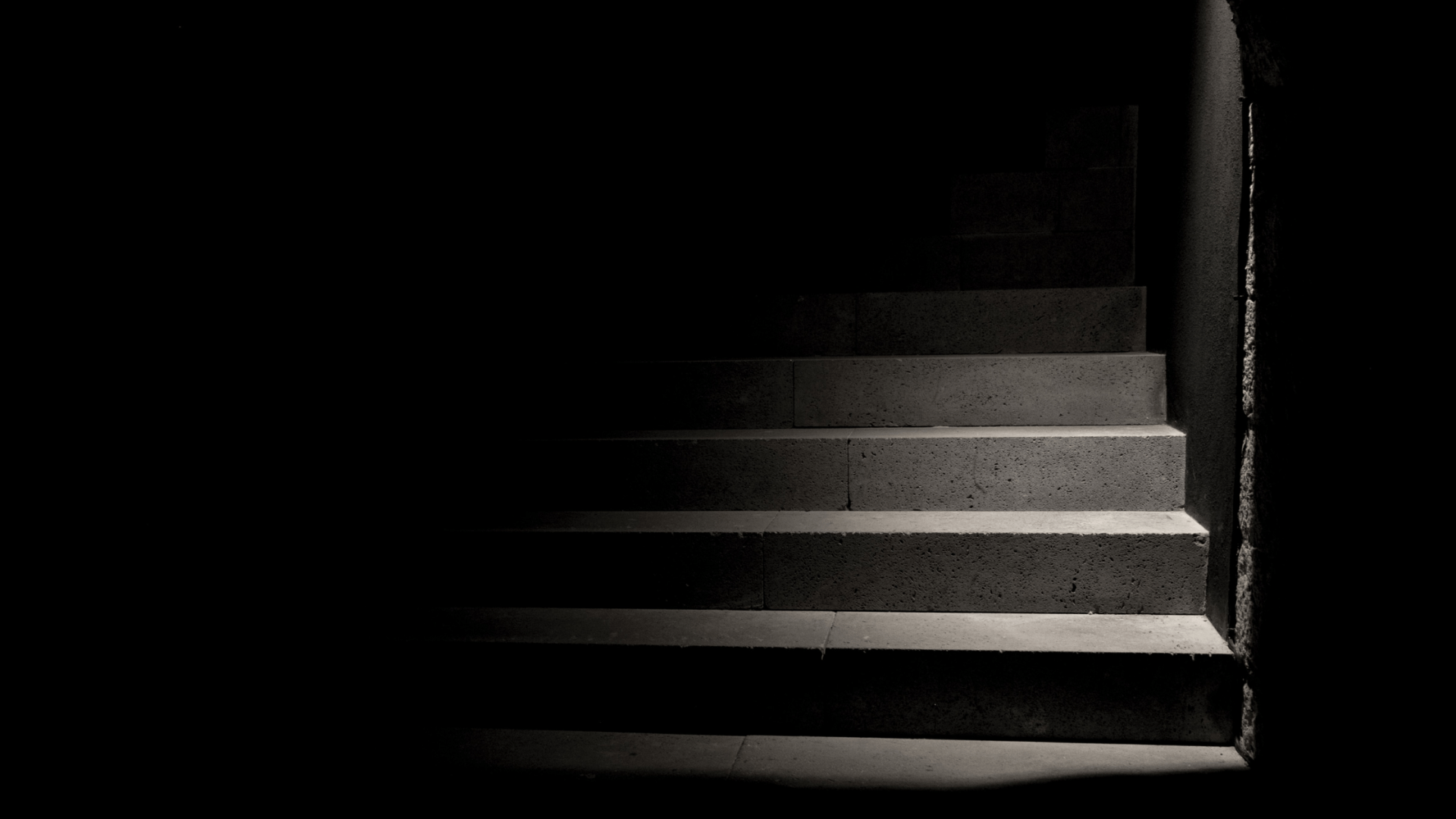 Light falling on grey cement staircase, going upwards in dark black hallway.