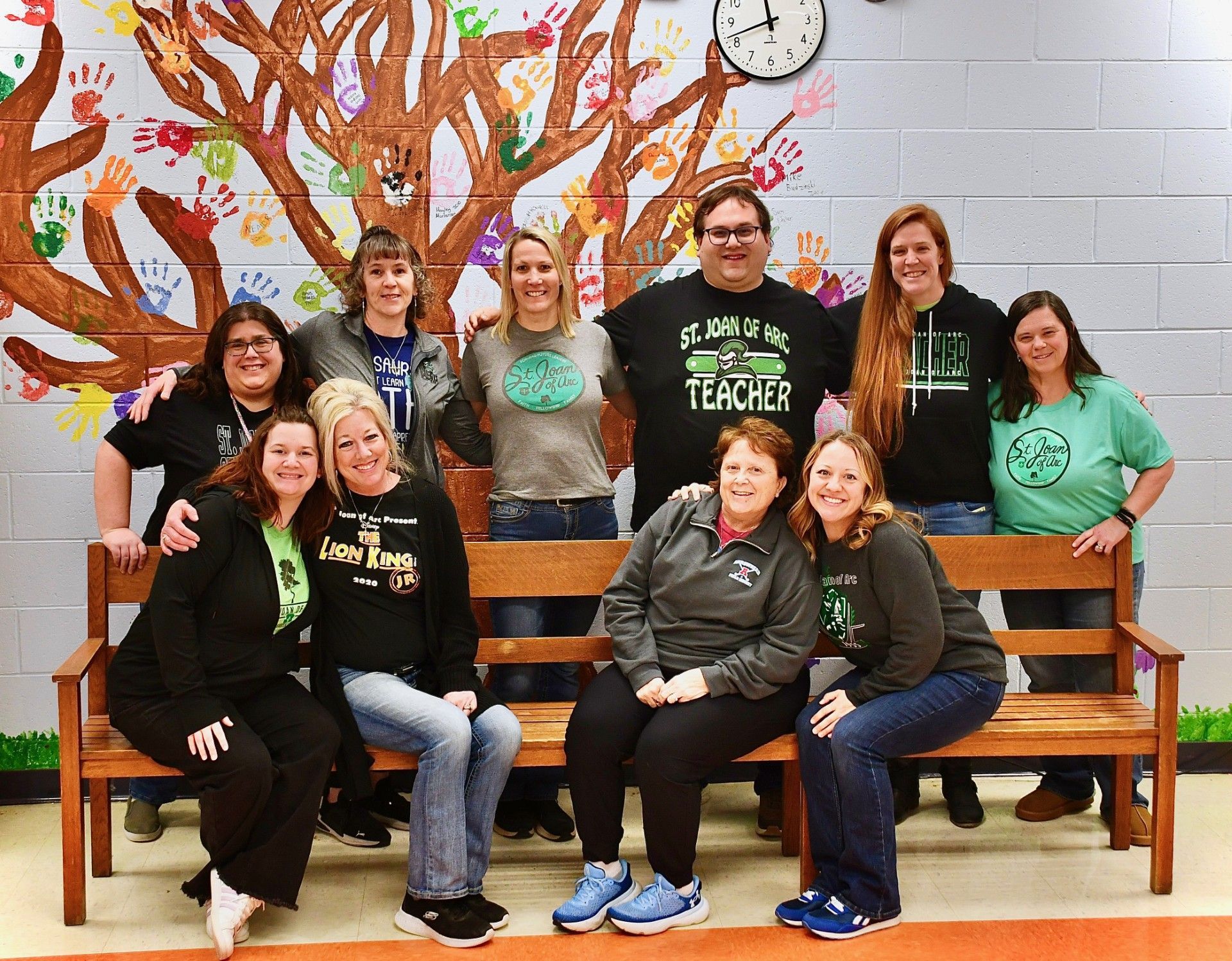 Group of teachers posing on a bench in front of a painted tree mural. Many wearing green shirts.