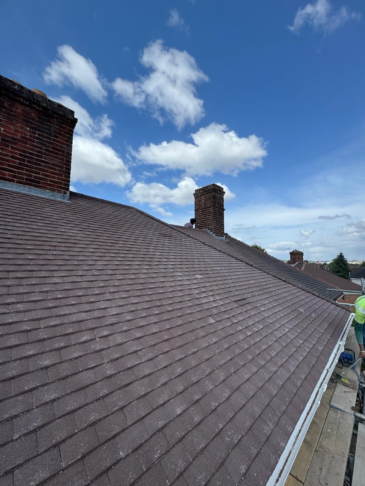 View of a dark, textured tiled roof with two brick chimneys against a blue sky with white clouds.