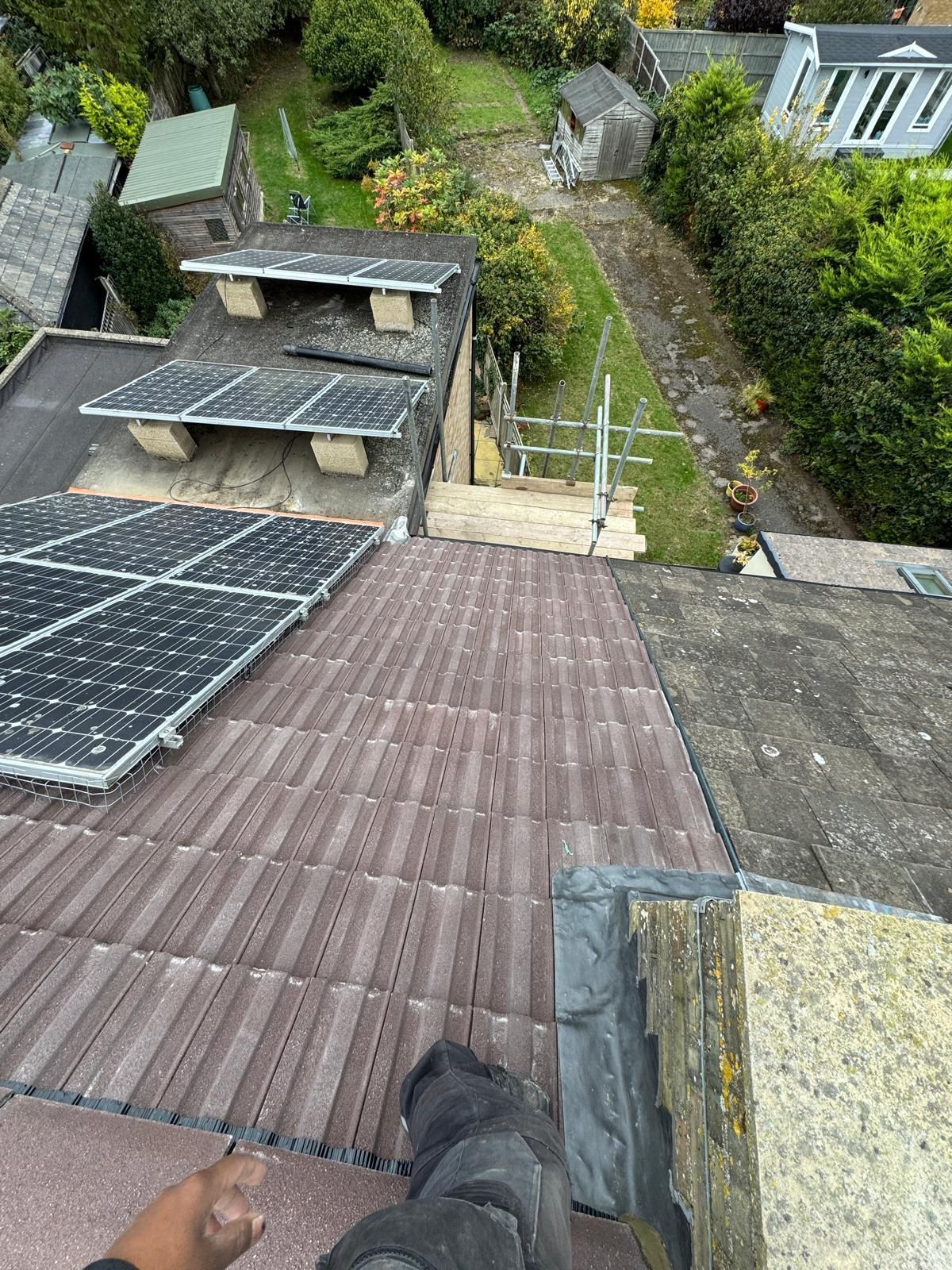 A view from a slanted rooftop showing solar panels, tiled roofing, a lower roof section, and a garden with sheds.