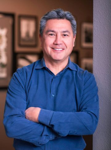 A man in a blue shirt is standing with his arms crossed and smiling.
