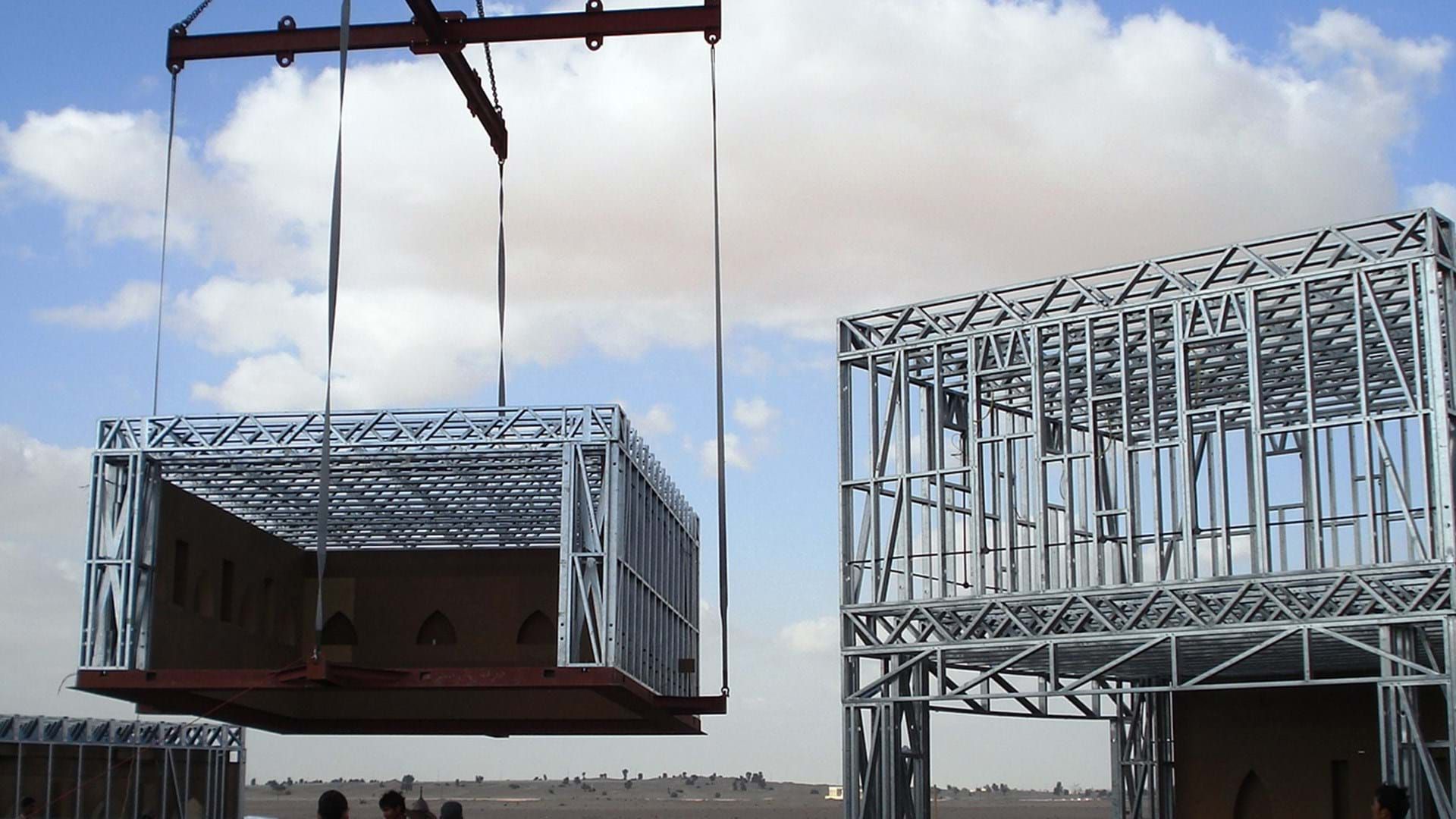 A crane lifts a prefabricated metal building module into position next to an existing steel-framed structure.