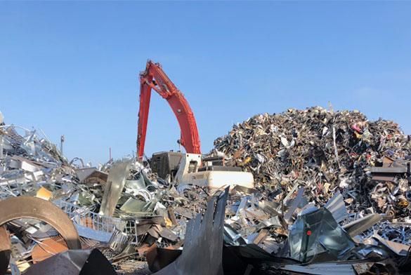 A red industrial excavator stands among large piles of scrap metal under a clear blue sky.