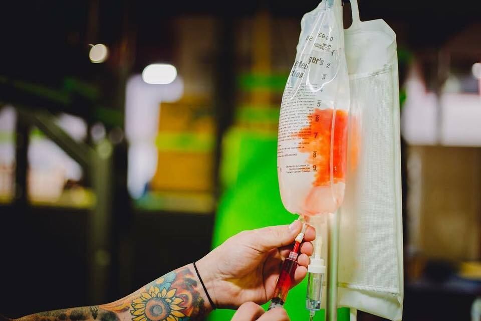 Person with tattoo adjusts IV drip bag with orange liquid.