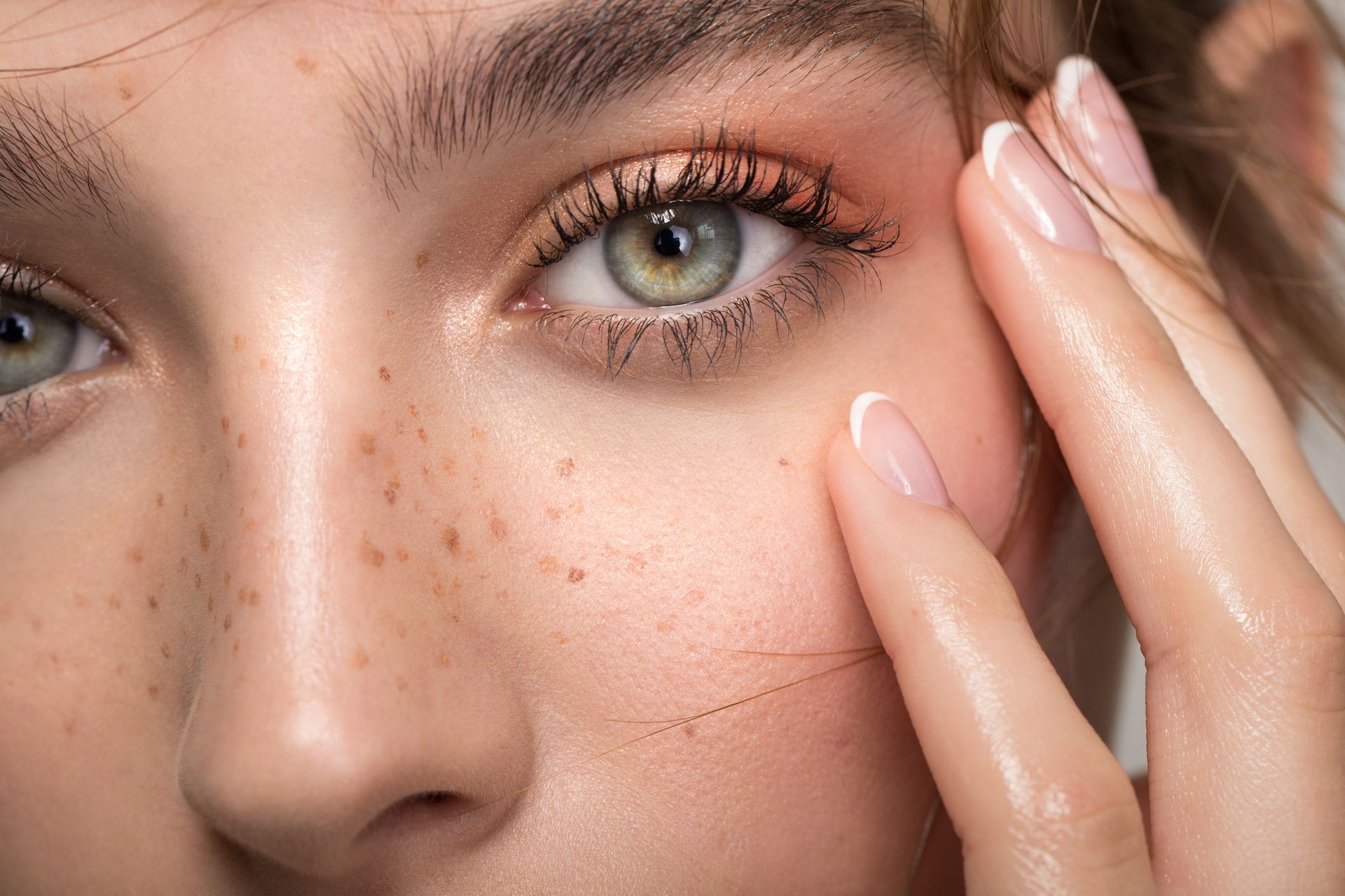 Close-up of a person's face with light skin, freckles, and green eyes, touching their cheek; they appear to be smiling.