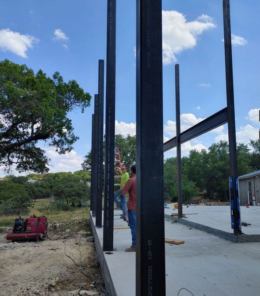 Construction workers erect steel beams on a concrete foundation. Blue sky and trees in the background.