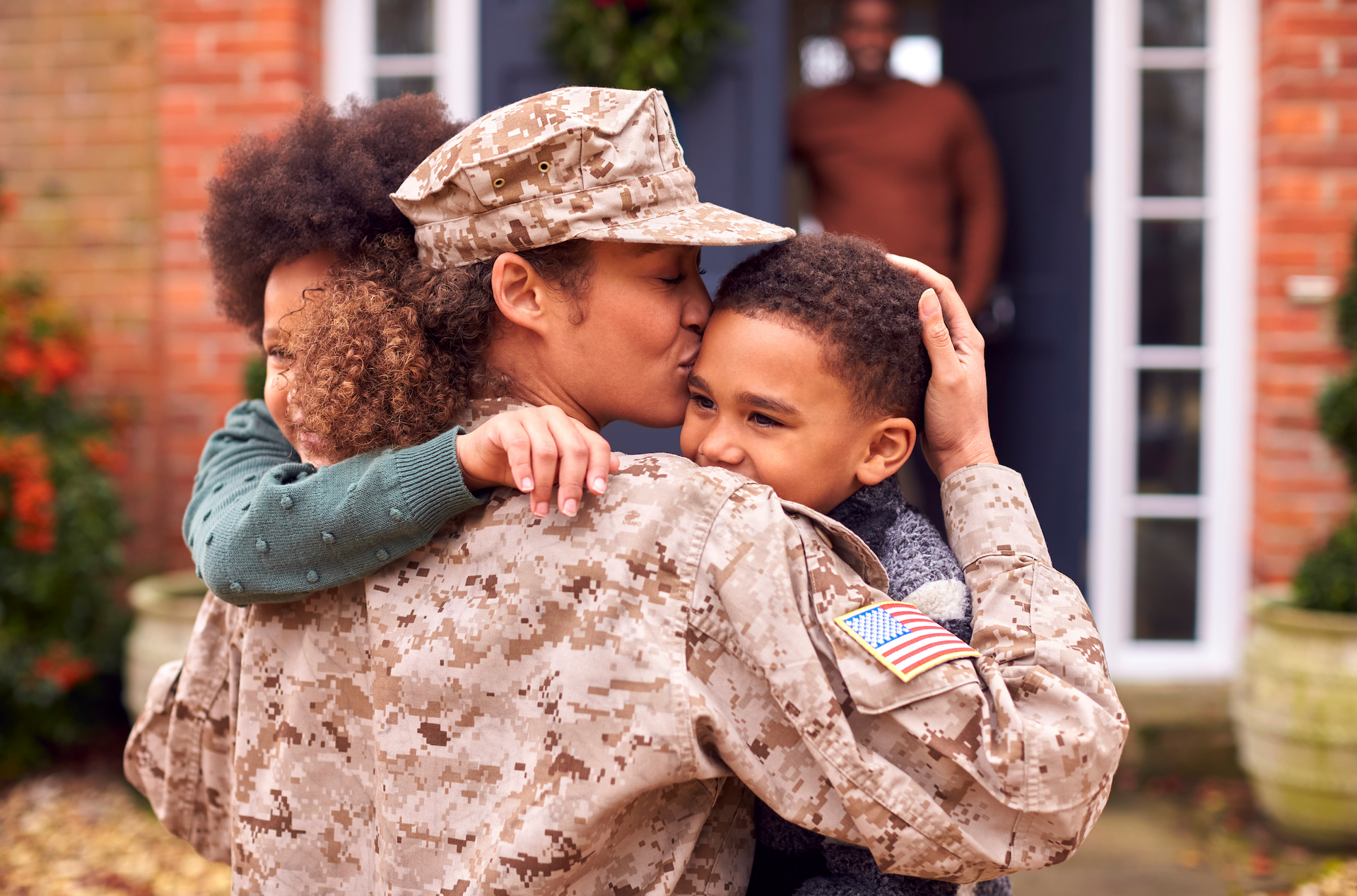 A woman in a military uniform is hugging a young boy in front of a house.