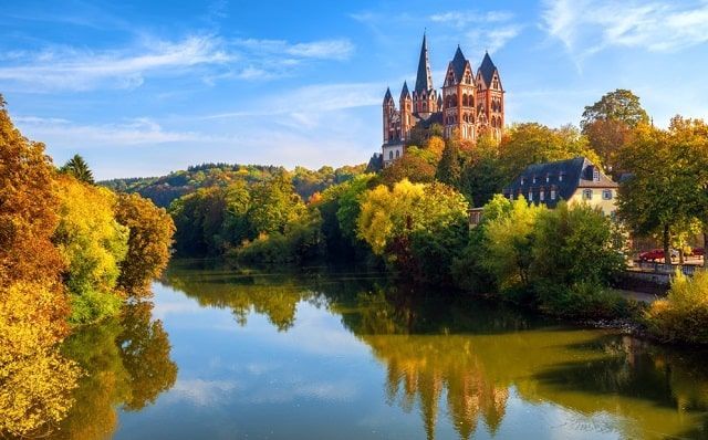 Een groot kasteel ligt op de top van een heuvel met uitzicht op een rivier, omringd door bomen.