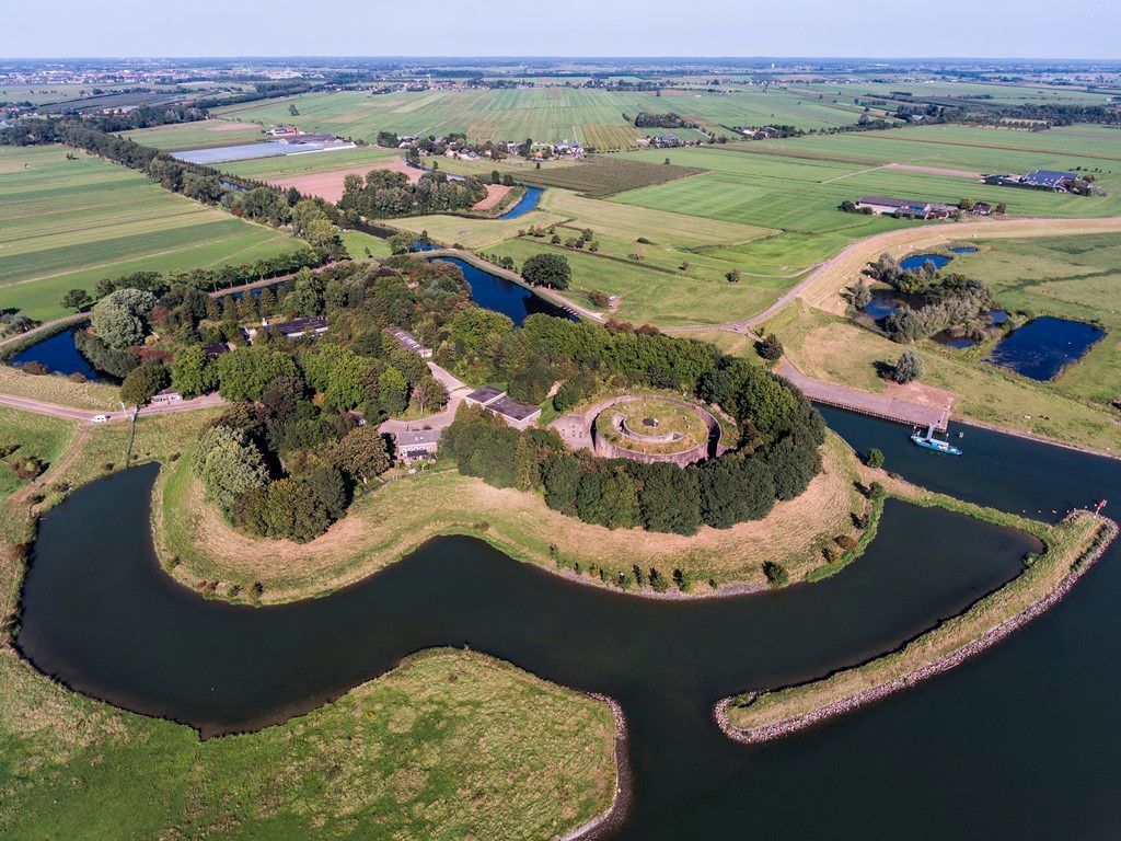 Een luchtfoto van een klein eiland in het midden van een rivier, omgeven door velden en bomen.