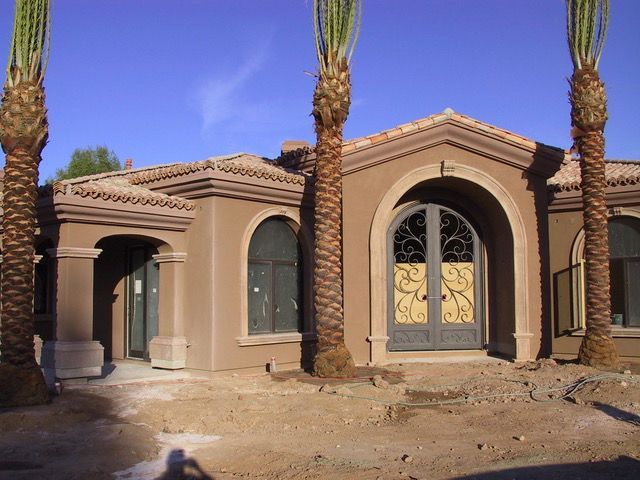Tan stucco home with arched doorway and ornate iron gate, flanked by palm trees.