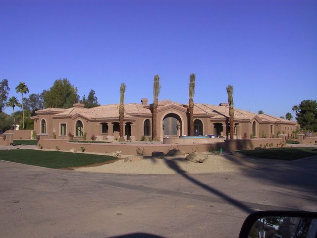 Large, single-story, stucco house with a tiled roof, arched entryway, and palm trees in front.