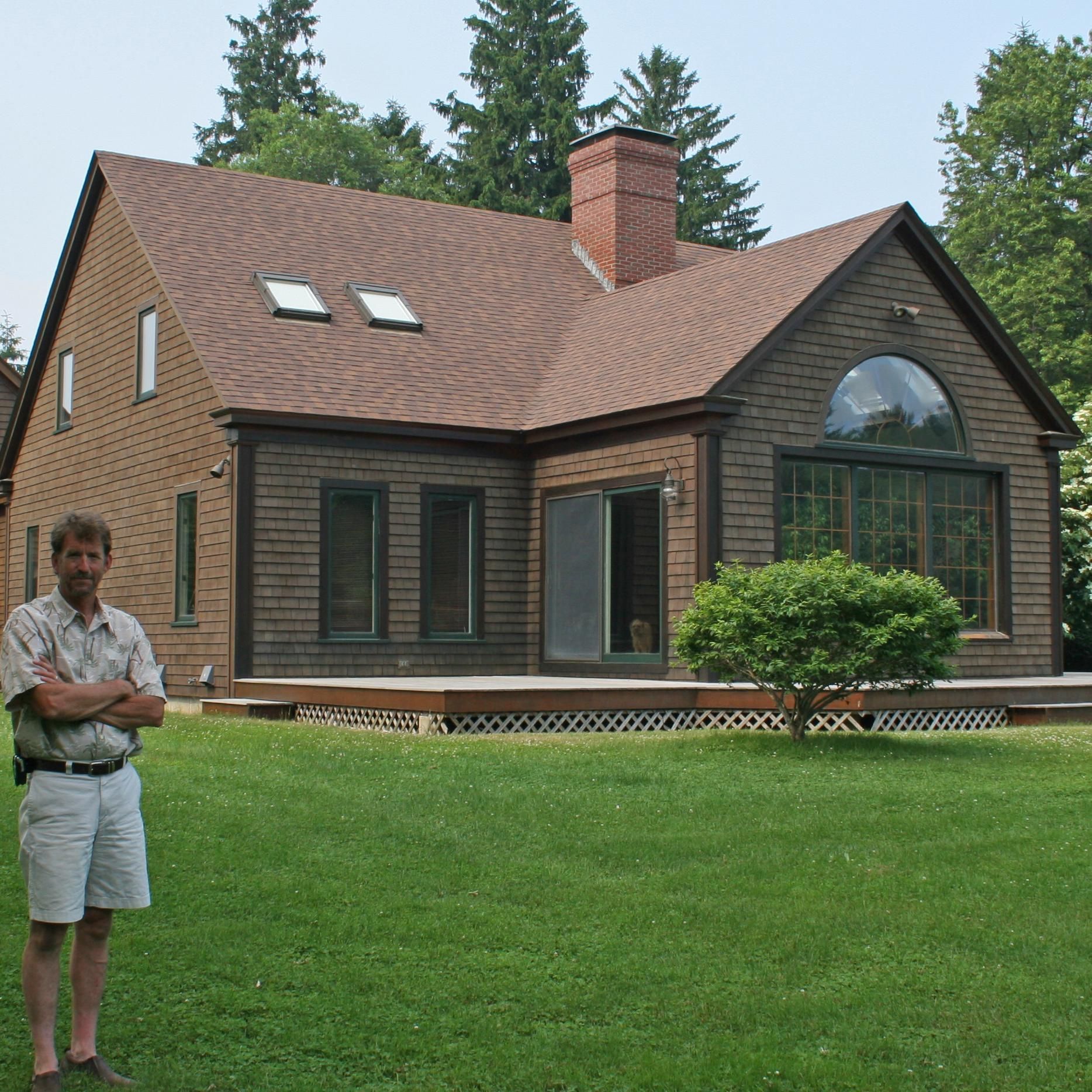Man standing in front of a brick house with a brown roof and chimney.