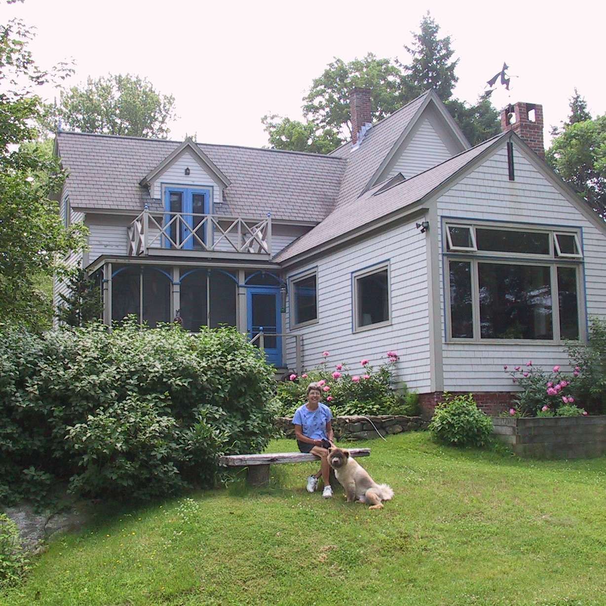 A woman sits on a bench next to a dog in front of a house