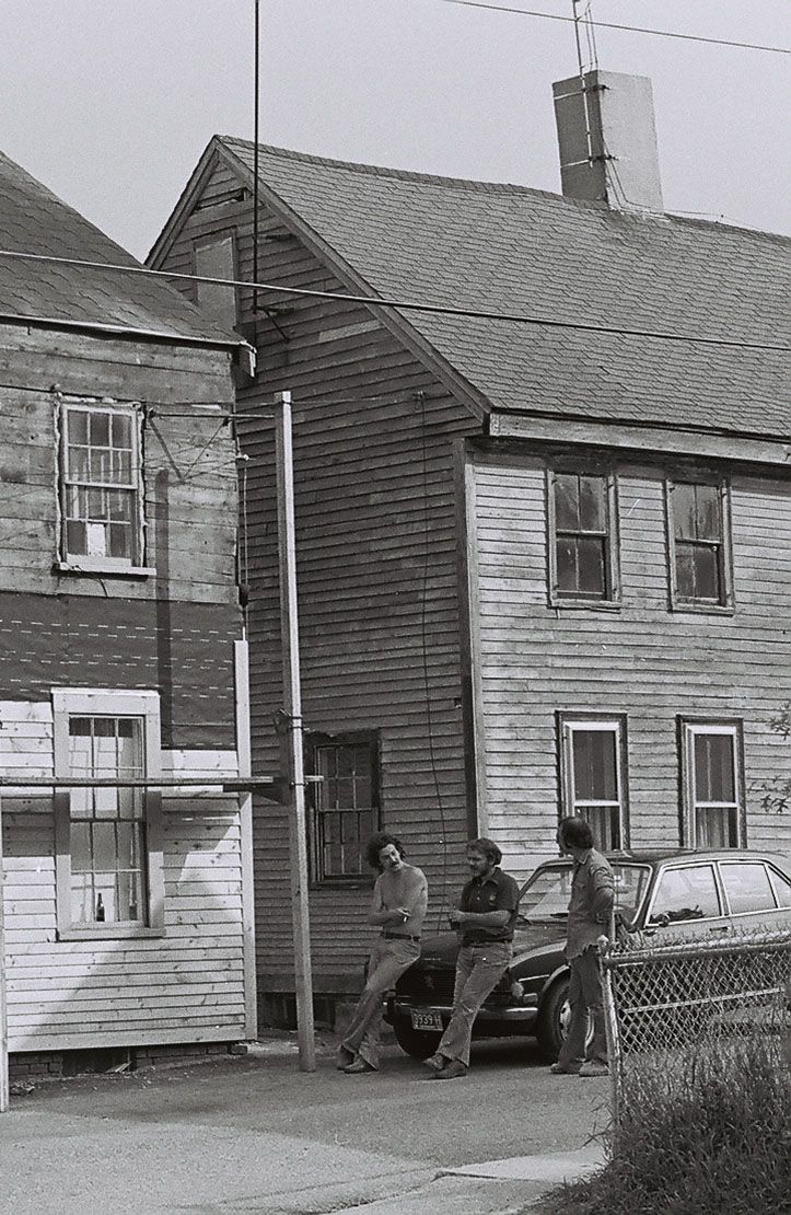 A black and white photo of two men sitting on a car in front of a wooden house