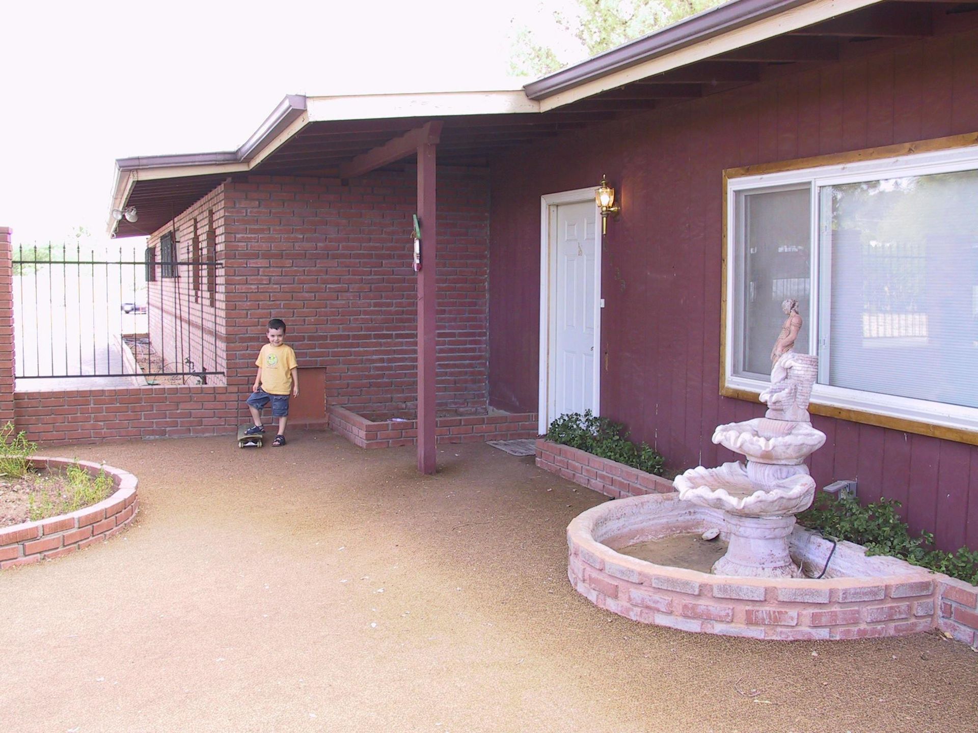 A young boy is standing in front of a red house with a fountain in front of it.