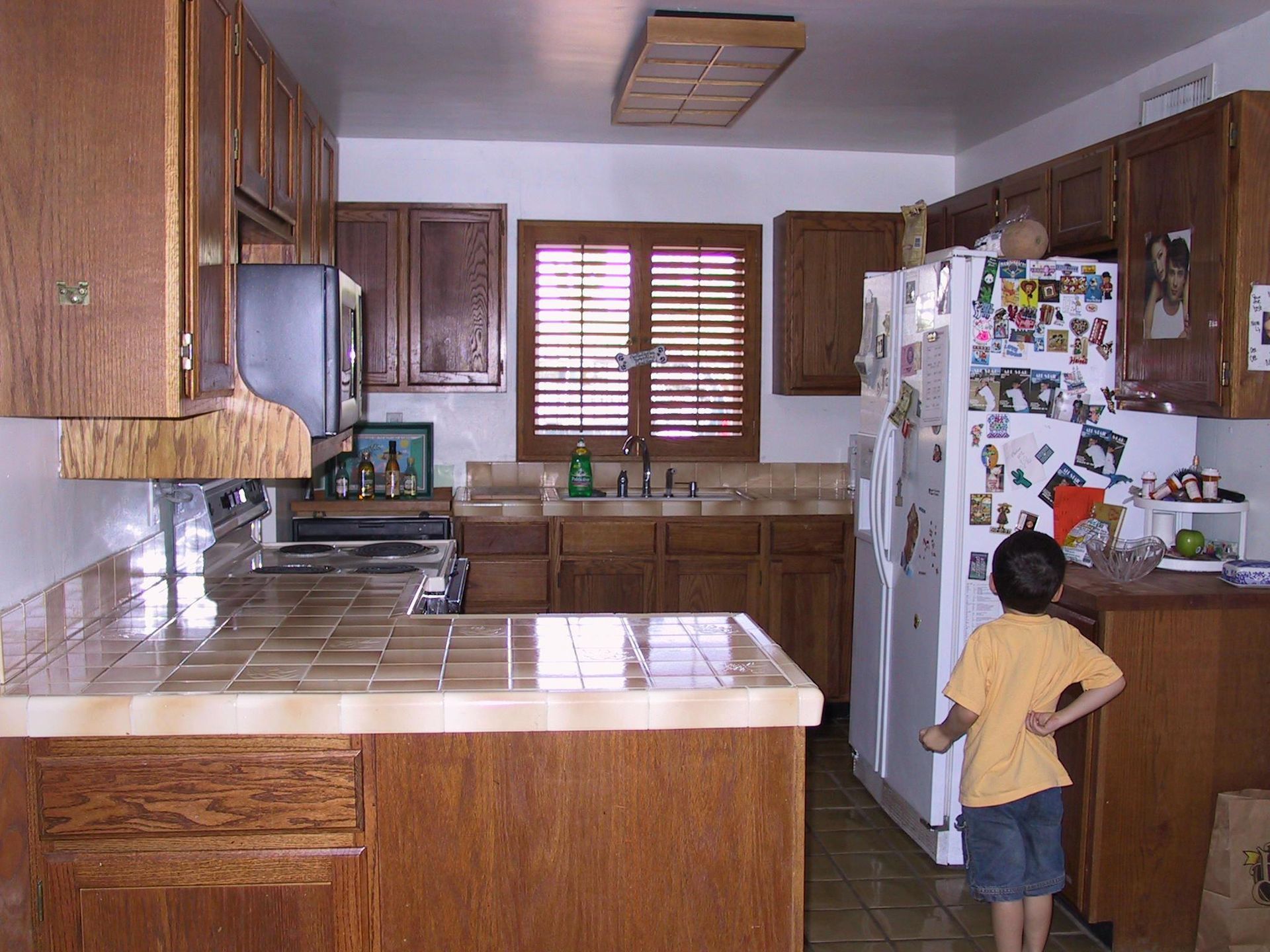 A boy stands in a kitchen looking at a refrigerator with stickers on it