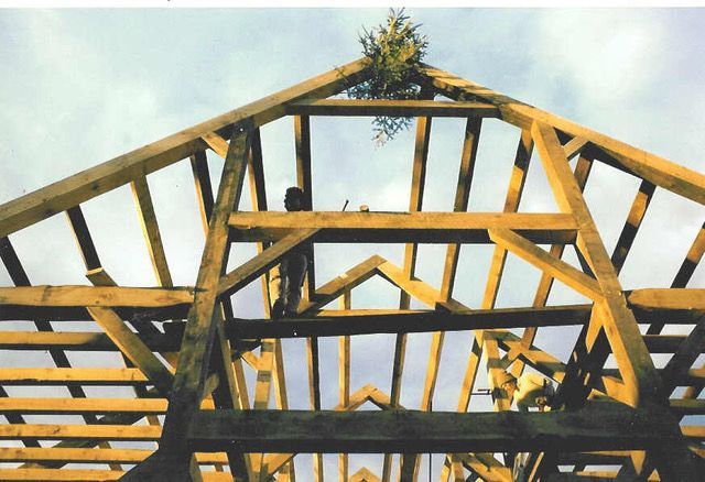 Wooden building frame under construction with blue sky, topped with greenery.