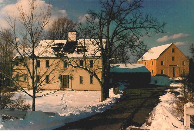 Snowy scene of a light-colored house and yellow barn. Bare trees and driveway lead to the structures.
