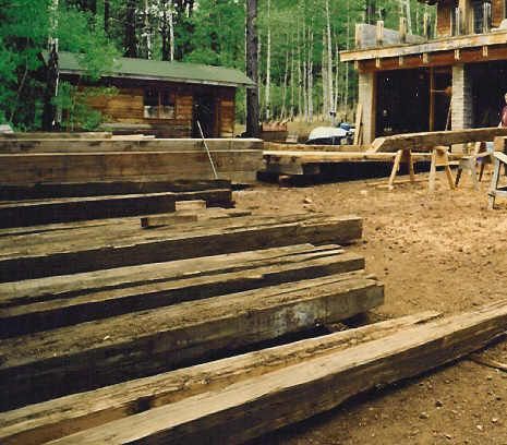 Wooden beams stacked outdoors at a construction site near a cabin and a partially built structure.