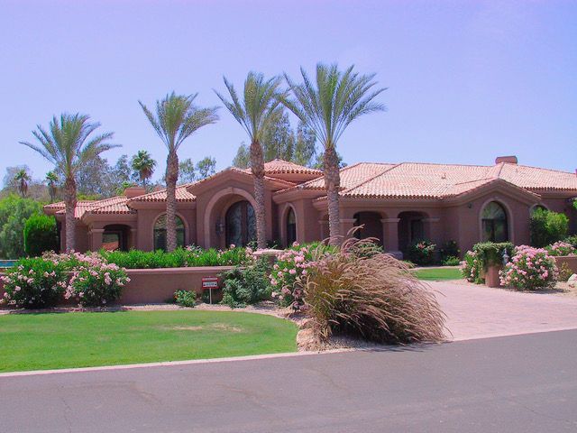 A terracotta-colored house with a tiled roof, palm trees, and manicured landscaping under a blue sky.