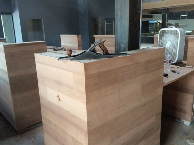 Wooden cabinets being constructed in a room; a hand plane rests on a cabinet. A fan sits nearby.