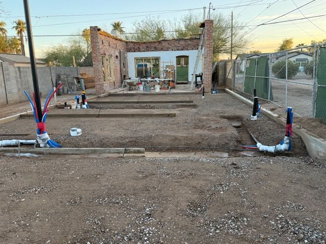 Construction site with stepped dirt terraces, exposed pipes, and a small unfinished building in the background
