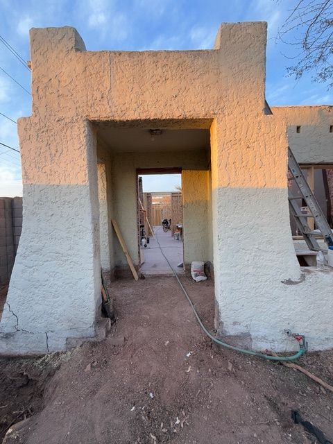 Unfinished adobe doorway under construction, with dirt ground, hose, and ladder on the right.