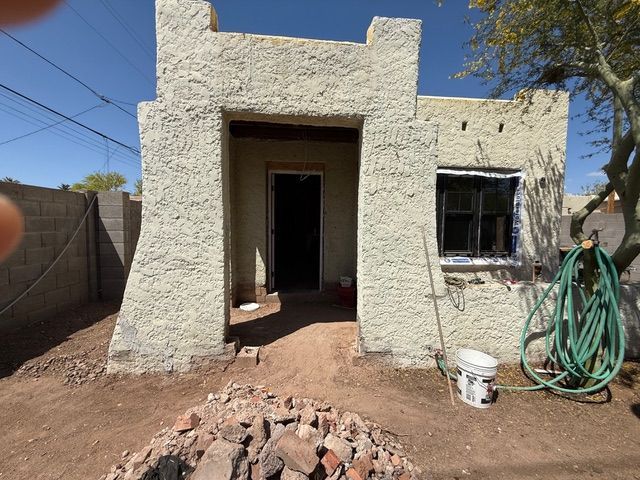 Small stucco house with open doorway, front window, hose, and rubble in the yard under a clear sky.