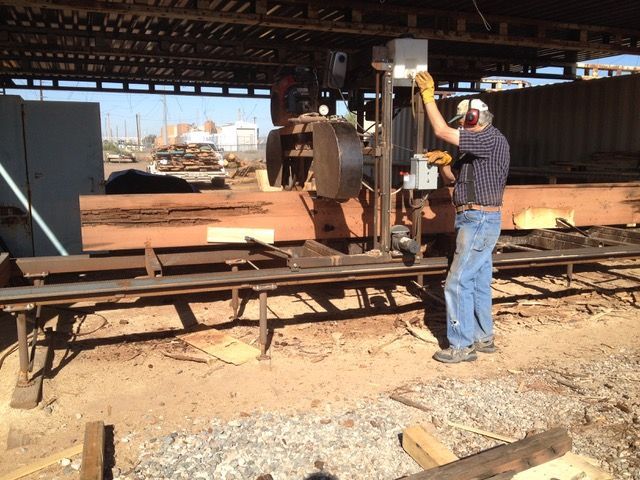 Man operating a large industrial sawmill, cutting lumber outdoors under a shed roof.