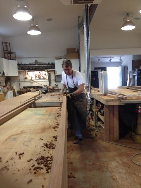 Man planing wood at a workbench in a workshop, sawdust visible, wearing an apron.