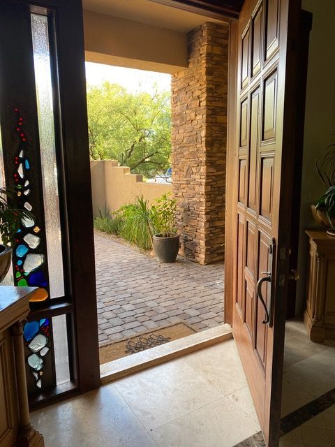 Wooden front door open to a brick patio, stone column, and greenery.