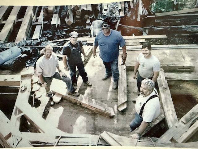 Five construction workers standing and sitting amid wood beams, looking at the camera.