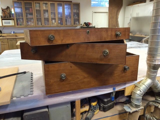 Three wooden dresser drawers, partially open, with brass knobs, on a workbench in a workshop.