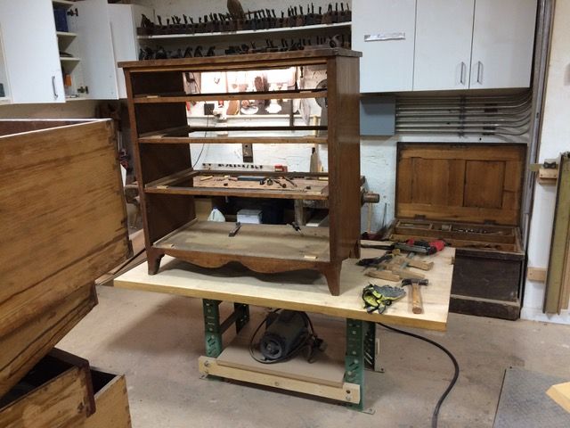 A wooden cabinet in pieces on a workbench in a workshop. Tools and other woodworking materials are nearby.