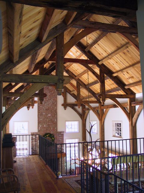 Interior view of a timber frame structure with wooden beams and a brick chimney. A second story railing is visible.